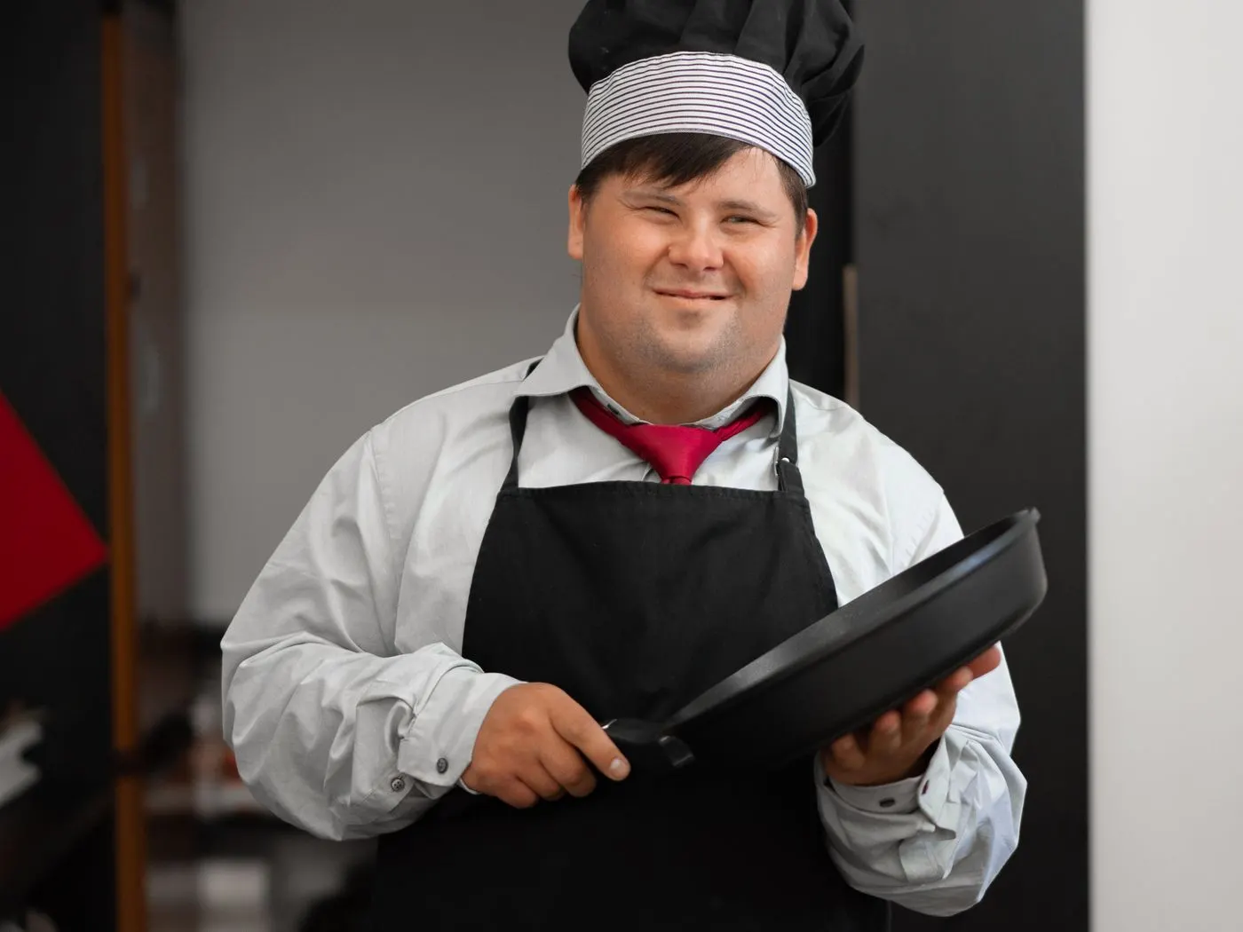 A man with Down syndrome in a chef's hat and apron, holding a frying pan and smiling